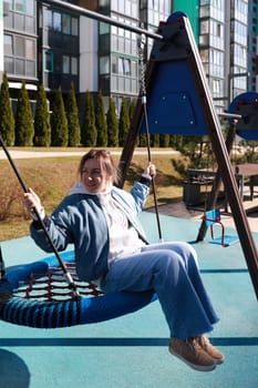 A young woman swings joyfully at a playground located in an urban area. The sunny weather enhances her cheerful expression as she enjoys her time outdoors.
