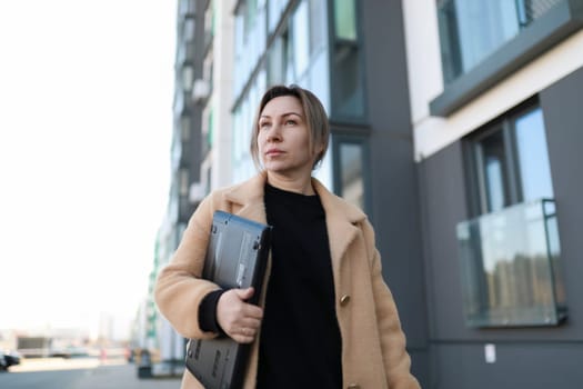 A woman in a beige coat strolls down a city street carrying a laptop. The modern building behind her reflects the bright skyline, showcasing urban life.