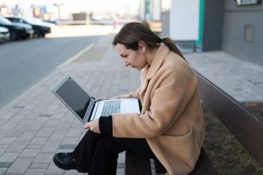 A woman sits on a wooden bench in a city, intently typing on her laptop. The surrounding area includes parked cars and a sidewalk, creating a lively urban atmosphere.
