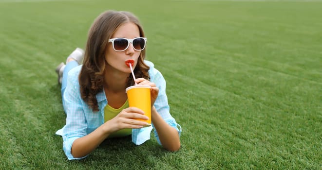 Happy modern young woman with cup of coffee, girl drinking fresh juice lying on green grass in summer park