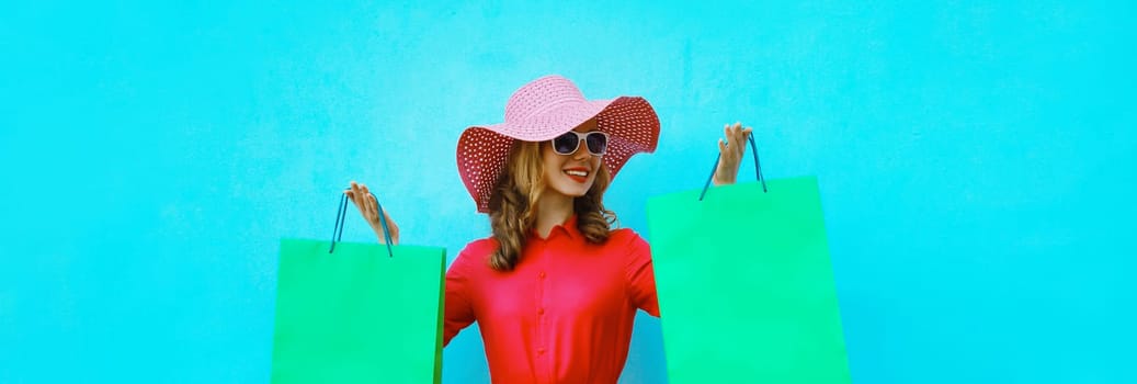 Beautiful happy smiling young woman model posing with colorful shopping bags in summer straw tourist hat, pink dress on blue studio background