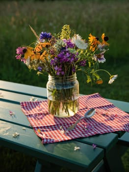 A jar filled with vibrant wildflowers is displayed on a checkered cloth on a green table. The warm golden hour sunlight highlights the flowers and lush greenery in the background.