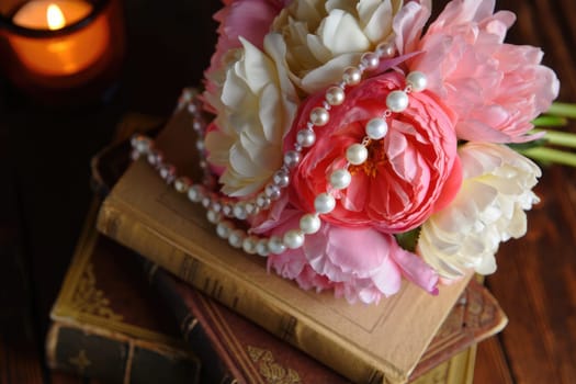 A beautiful bouquet of pink and white flowers rests on a stack of vintage books. A lustrous pearl necklace adorns the flowers, creating a warm, inviting ambiance with a candle in the background.