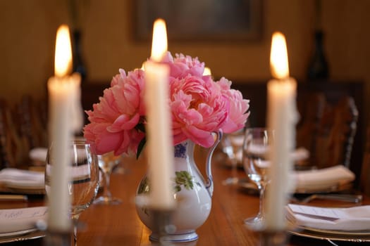 A beautifully arranged dining table features a vase of pink peonies surrounded by lit candles. Glassware is placed neatly, creating a warm and inviting atmosphere for a special gathering.