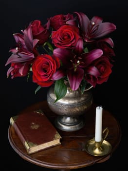 A beautiful arrangement of red roses and lilies showcased in a silver vase on a wooden table. Next to it, a lit candle and an antique book add to the charming ambiance.