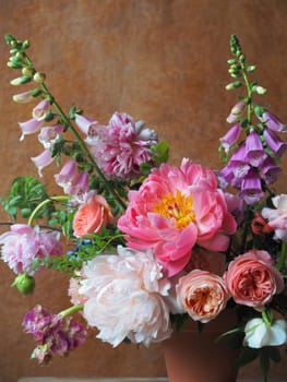 A stunning floral arrangement showcases an array of colorful flowers, including peonies and foxgloves. The vibrant hues are set against a warm, textured background, creating a cheerful atmosphere.