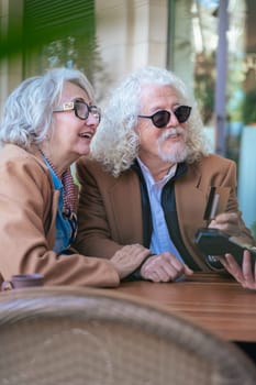 Joyful caucasian senior couple at an outdoor cafe making a card payment. Both wearing trendy outfits and sunglasses. Ideal for retirement, finance, lifestyle, and senior couple relationship concepts.