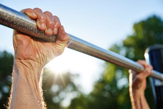 An elderly man is gripping a horizontal bar, performing pull ups in an outdoor park setting during a bright morning. The sun is shining, highlighting his determined effort.