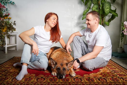 A couple relaxes on a rug with their German Shepherd, sharing a moment of joy in a warm indoor environment.