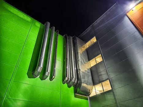 Urban building features a striking green facade and metallic ducts illuminated at night, highlighting modern design.