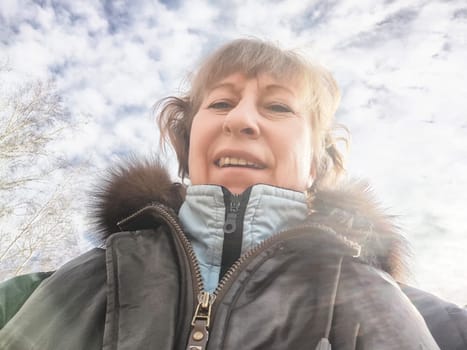 A woman in winter attire smiles while taking a selfie against a backdrop of clouds and blue sky.