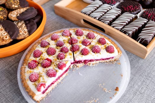 Raspberry tart with assorted cookies and chocolate biscuit display on grey tablecloth.