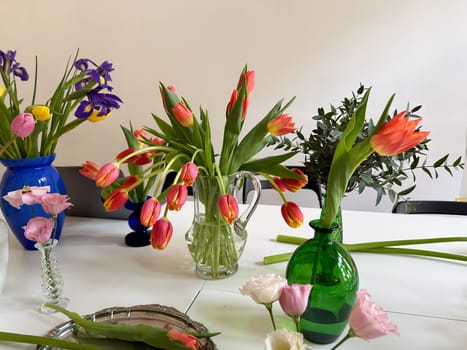 Mixed flower arrangements in colorful vases on white table in bright interior. Spring mood and creative floral styling at home.