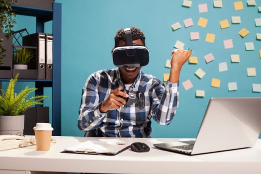 Cheerful male entrepreneur wearing VR goggles, celebrating with his fist raised and game controller in hand. Manager engaged in virtual reality with headset, interacting with a 3D gaming simulation.
