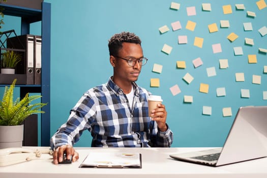 African american startup employee focused on laptop screen, analyzing business metrics at his desk. Marketing specialist holds a mouse and coffee cup browsing online, reviewing research data.