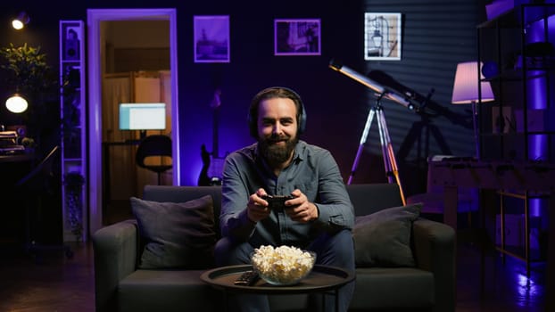 Cheerful gamer wearing headphones, chatting with teammates while playing videogames. Jolly person at home enjoying defeating enemies in game, talking with friends through headset mic, camera B