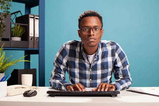 Serious male manager leaning at his office desk, typing on his computer keyboard, focused on updating company information. POV of african american analyst closely reviewing startup project results.