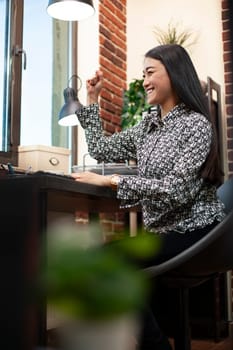 Asian woman celebrates at desk with her fist raised, after reading positive business outcomes on her computer. Excited female manager smiles, reflecting on success of company project.