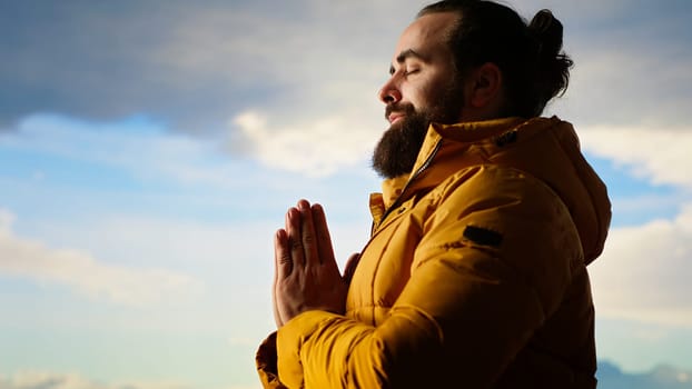 Adventure seeker doing a meditation pose on top of the mountain, successfully climbing the hill and enjoying the scenic landscape. Young calm zen traveler hiking and meditating. Camera B.