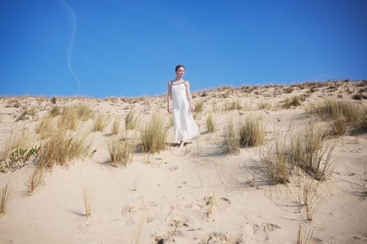 A barefoot woman in a flowing white dress walks peacefully down a sandy dune. The bright blue sky and dry beach grass create a serene, natural setting full of open space and calm.