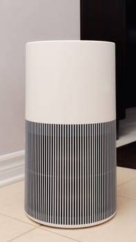 Tall round air purifier with a black top fan and a white body on a tile floor against the wall.
