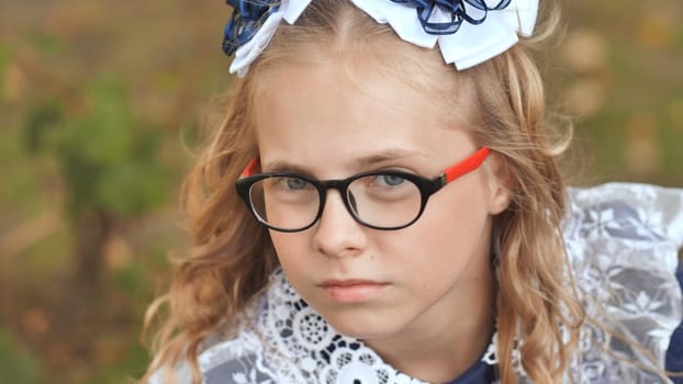 Schoolgirl wearing glasses, hair bows, posing seriously near outdoor setting with neutral background