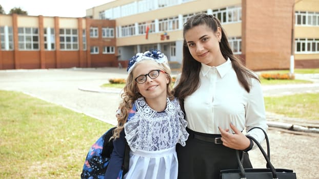 Schoolgirl wearing white dress and teacher smiling together in front of school building on their first day of school