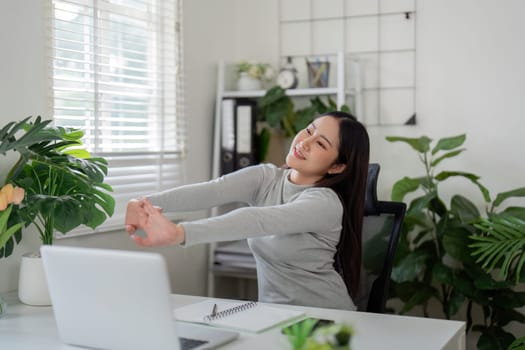 In her serene workspace, a young woman does a gentle stretch, promoting wellness and relaxation while working from home.