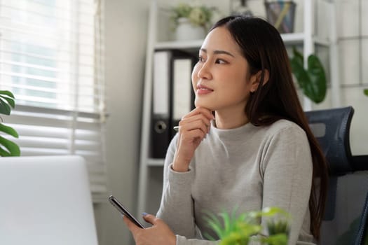 A young woman sits in her home office, holding a smartphone while deep in thought about her tasks. The image conveys concentration and the essence of remote work.