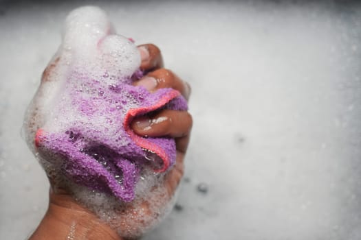 A hand squeezes a purple sponge, creating bubbles in soapy water.