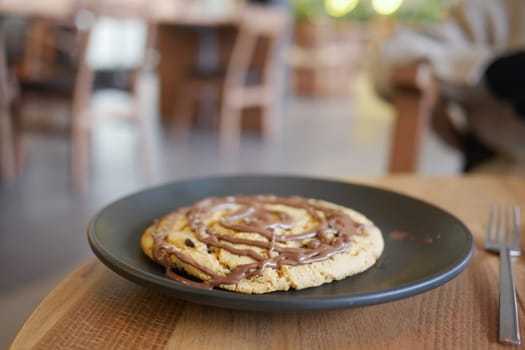 A large cookie topped with chocolate sits on a plate in a cozy cafe.