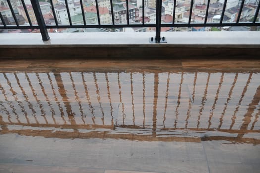 Raindrops create puddles on the balcony floor reflecting buildings.