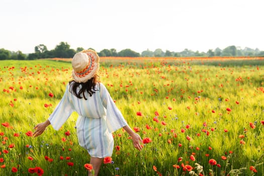 A girl in a straw hat, dress stands in a beautiful poppy field with red poppies. Sunrise, happiness, morning walk