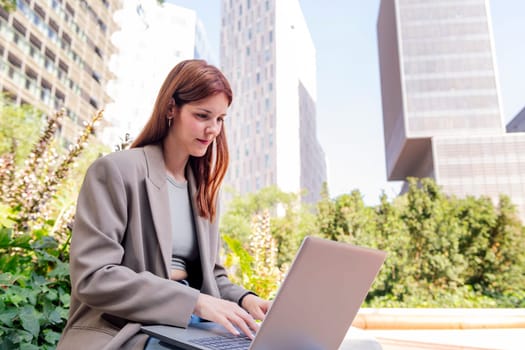 smiling young woman using a laptop sitting on a bench of a park near of office buildings, concept of business and technology for remote work