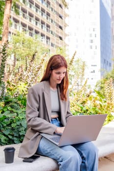 smiling young woman using a laptop sitting on a bench of a park near of office buildings, concept of business and technology for remote work