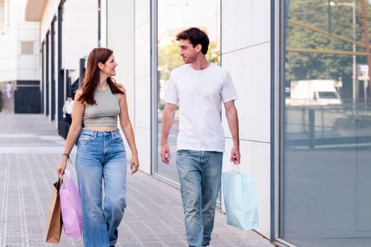 young adult man and a woman in casual clothes walking down a sidewalk near of a mall carrying shopping bags, concept of couple and urban lifestyle, copyspace for text