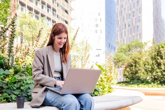 smiling young woman using a laptop sitting on a bench of a park near of office buildings, concept of business and technology for remote work