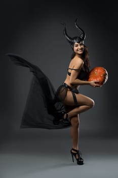 Full body side view of smiling female model wearing provocative black devil fairy with horns costume standing on one leg with pumpkin in hands