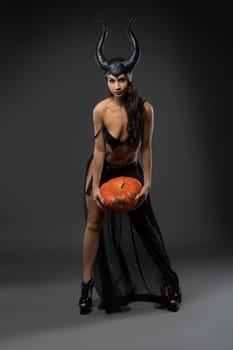 Full body of smiling female witch model wearing provocative black devil fairy with horns costume standing with pumpkin in hands