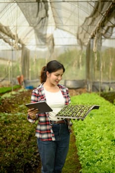 Smiling woman holding a tray of seedlings while checking data on digital tablet inside a greenhouse.