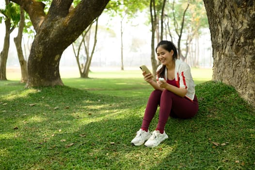 Young woman sitting under a tree holding wireless earbuds and using smartphone.