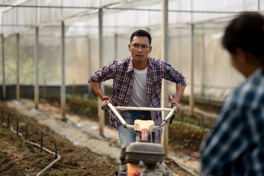 Male farmer in a plaid shirt using rotary tiller to prepare soil in a greenhouse.