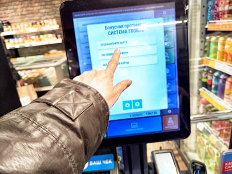 Kirov, Russia - February 14, 2025: A person uses a touchscreen menu to check options at a grocery store in the evening, surrounded by various product displays.