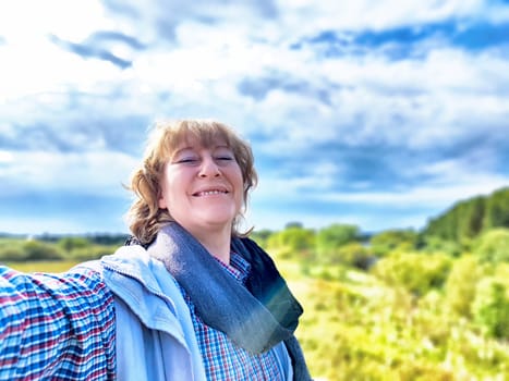 A middle-aged woman takes a cheerful selfie in a vibrant landscape, enjoying nature during her travel adventure.