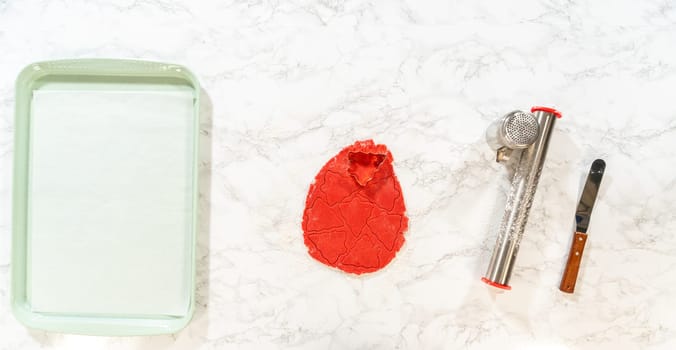 On a marble countertop, a smooth red ball of gingerbread dough is ready to be rolled. Next to the dough, a rolling pin, flour sifter, and offset spatula are arranged, creating a clean and organized baking setup for making Red Gingerbread Cookies.
