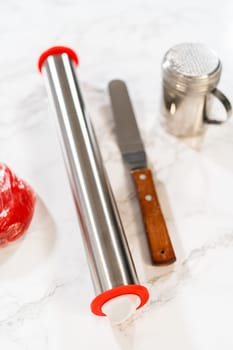 A collection of baking tools, including a rolling pin with red handles, a butter knife with a wooden handle, and a flour sifter, arranged on a marble countertop. These tools are essential for preparing Red Gingerbread Cookies.