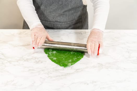 A close, eye-level view capturing hands using a rolling pin to flatten green dough on a marble surface. This step prepares the dough for cutting out shapes for the Green Gingerbread Cookies recipe.
