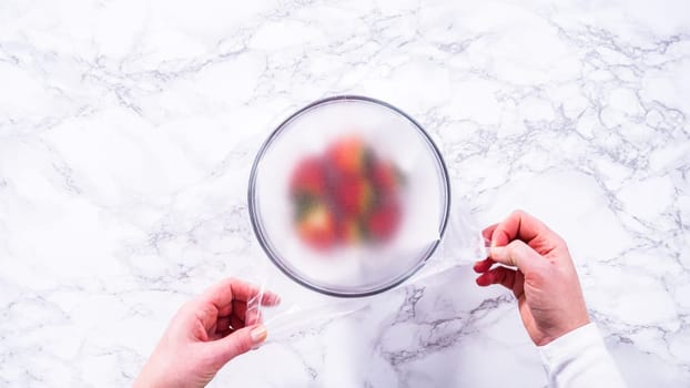 Flat lay. Strawberries, freshly washed and dried, are neatly stored in a glass bowl lined with a paper towel and securely covered with plastic wrap to maintain freshness.