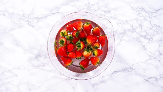 Flat lay. Ripe strawberries are submerged in water within a large glass mixing bowl, a step in washing the fruit to ensure cleanliness and longevity before storage or consumption.