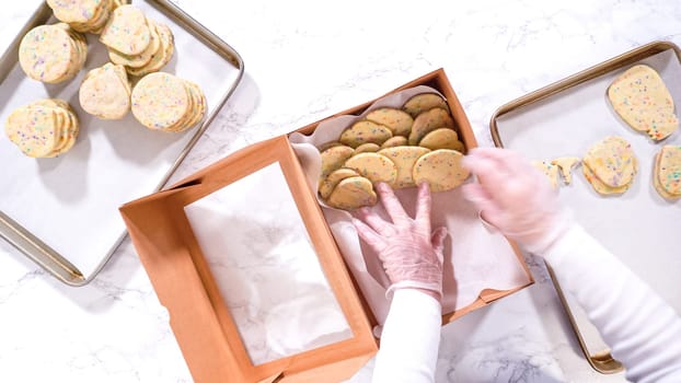 Flat lay. With precision, the woman is carefully arranging the sugar cookies, filled with dough-mixed sprinkles, into a rustic brown paper box.
